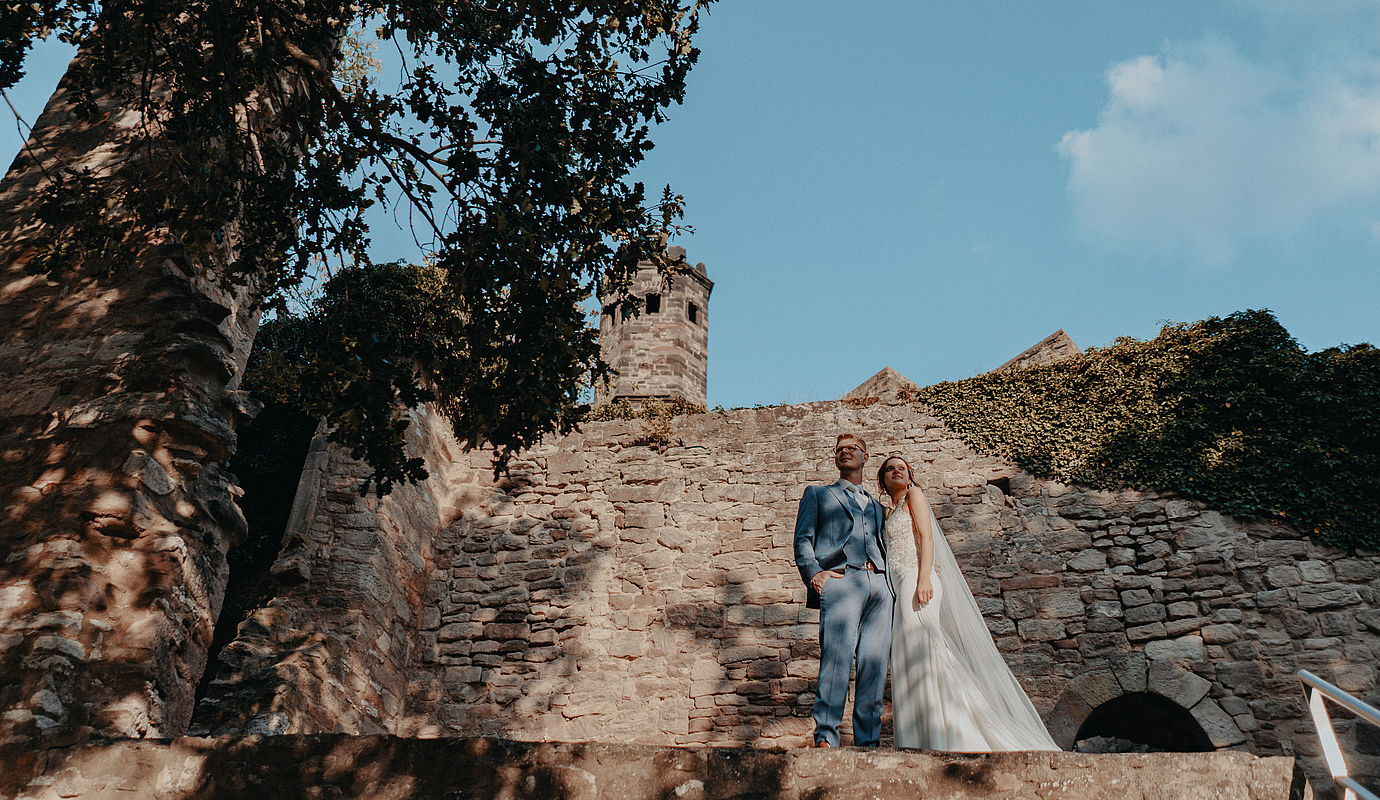 Braut und Bräutigam umarmt mit Blick in die Ferne stehen am Fuße der Burg bei ihrer Hochzeit in Niedersachsen