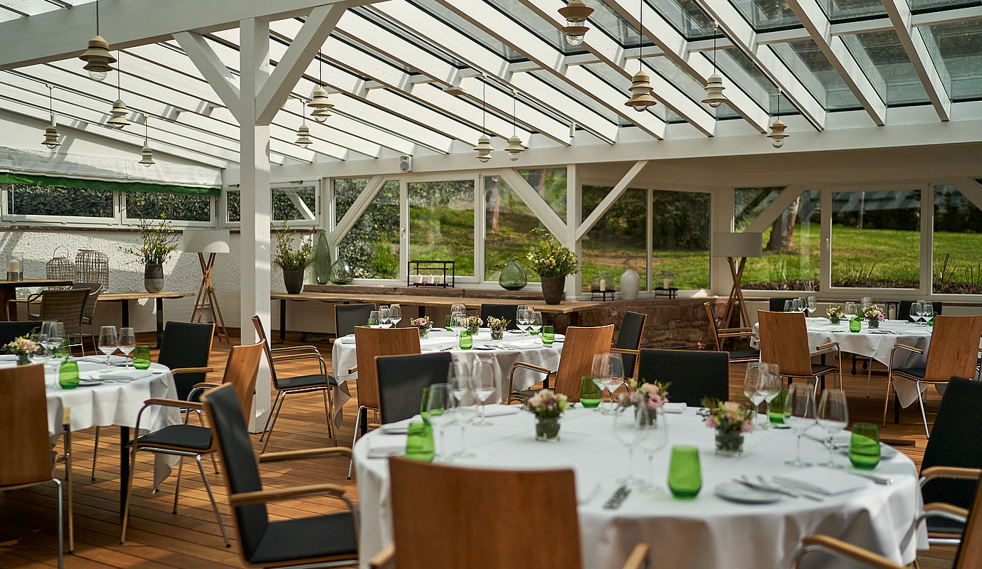 Stühle und runde Tische mit weißen Tischdecken und Blumenschmuck in einer Eventlocation in Niedersachsen mit großen Fenstern mit Gartenblick.