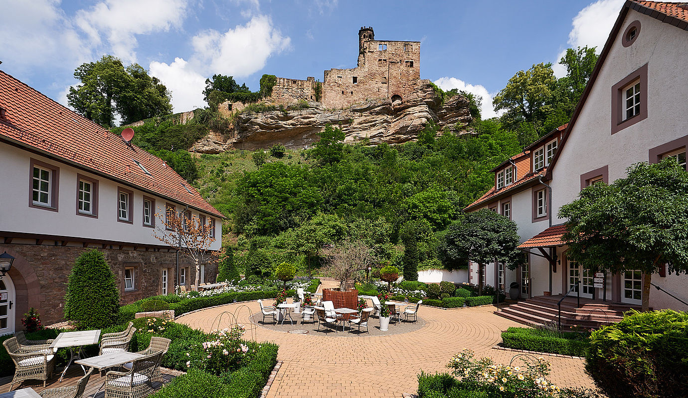 Idyllischer Innenhof eines Hotels mit Gartentischen und Blick auf eine mittelalterliche Burgruine.